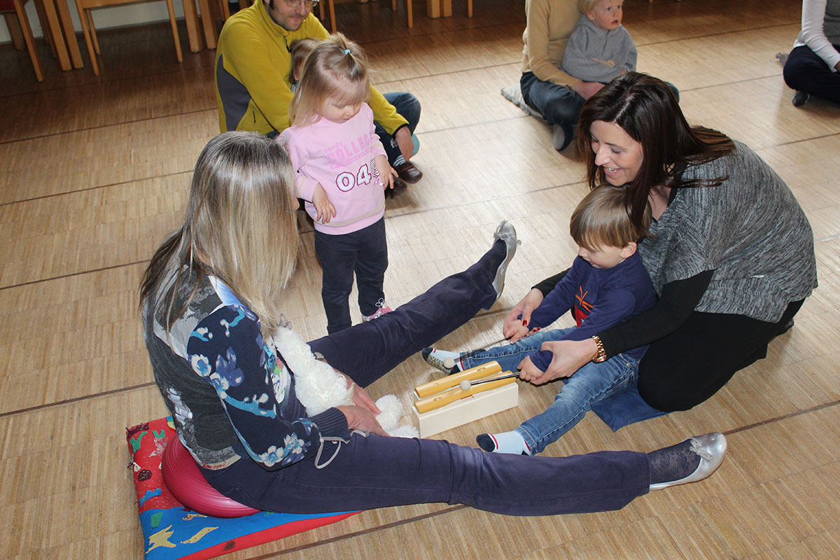 Musikgarten, ein Kind spielt zusammen mit seiner Mutter ein Orff-Instrument, ein anderes Kind hört zu